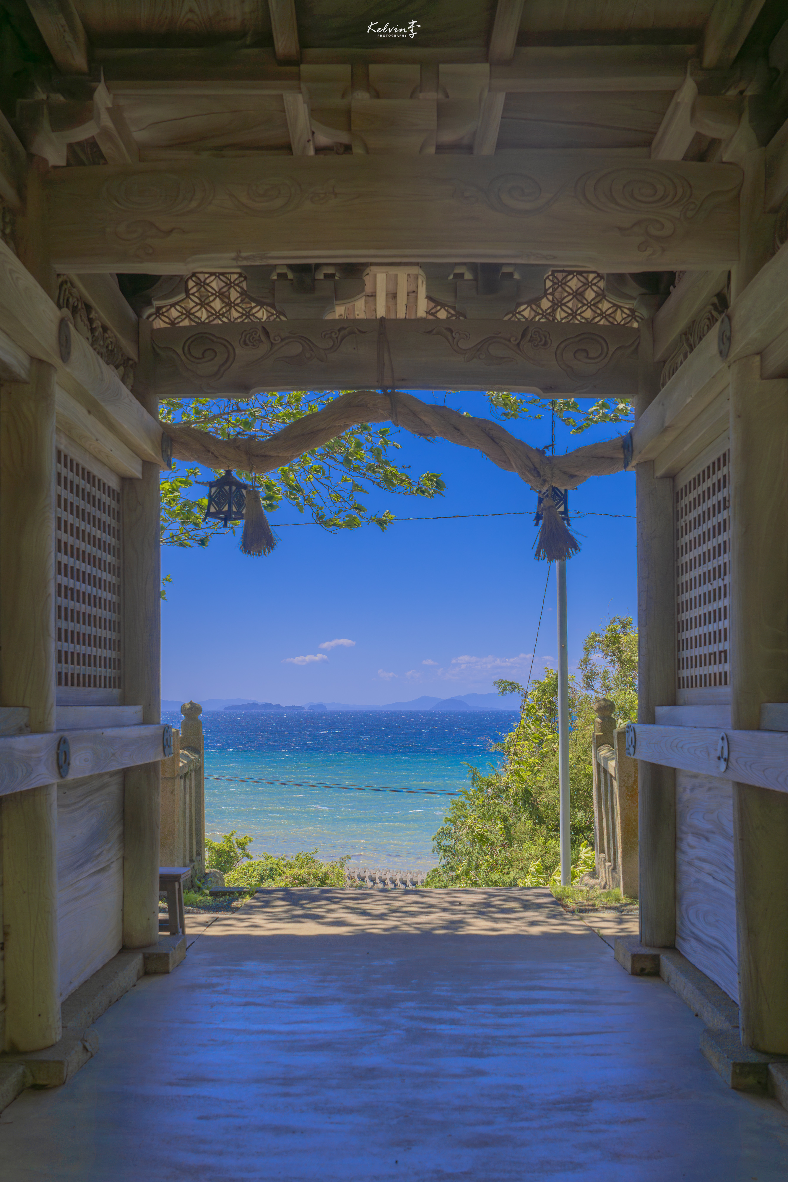 木造の門と注連縄の向こうに、青い空と海が広がる風景。