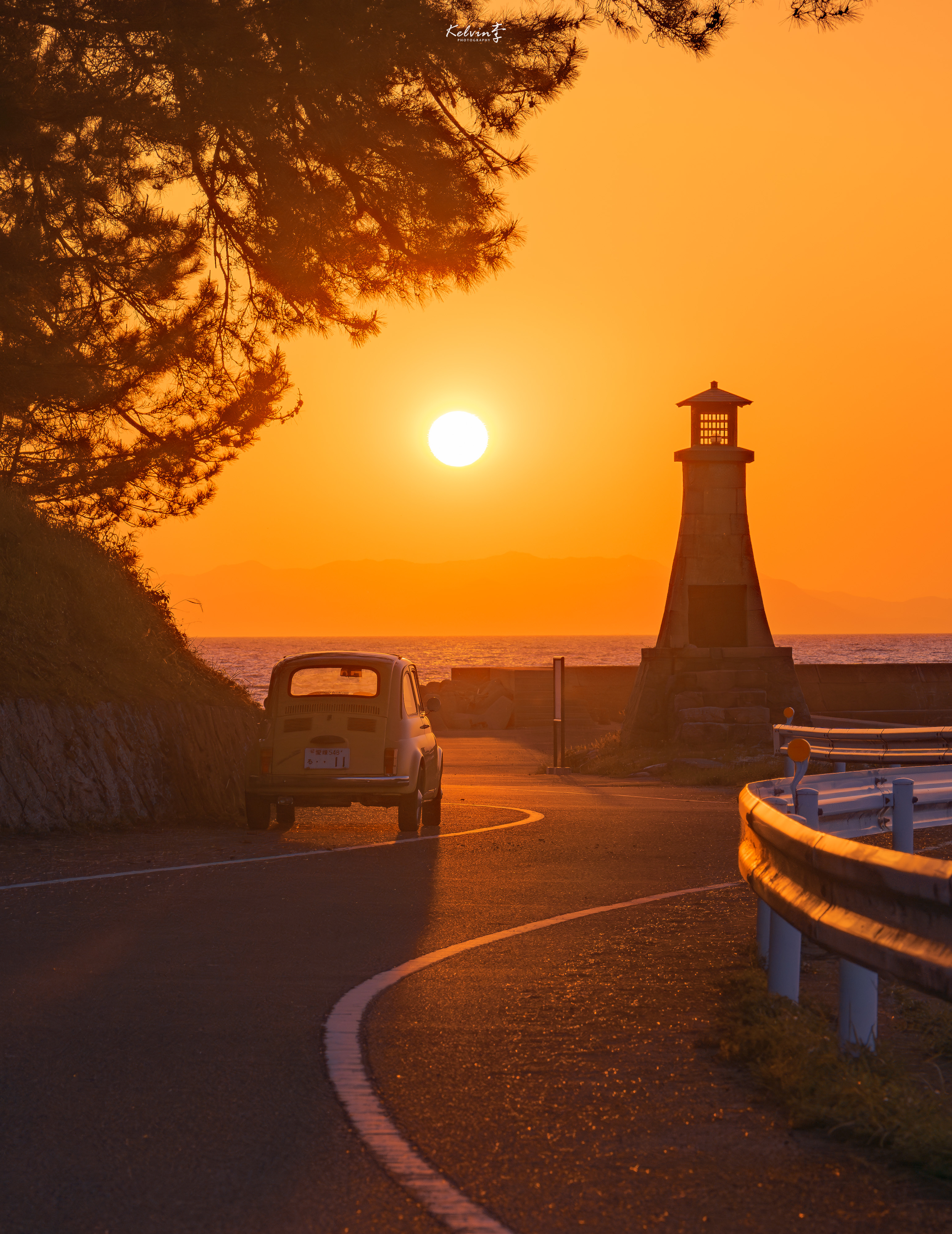 オレンジ色の夕日に染まる海辺の道と、車、石造りの灯台。